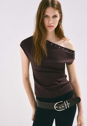 Young woman with long brown hair wearing an off-shoulder dark top with studs and a wide black belt on black pants, neutral background.