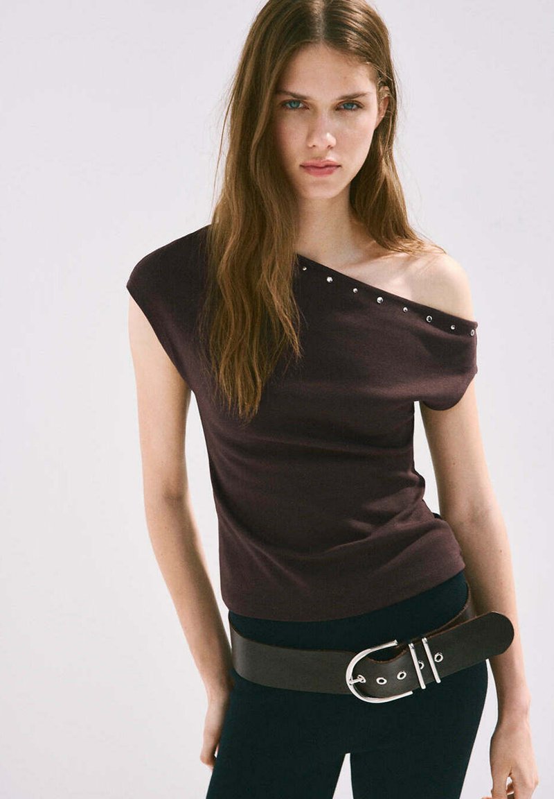 Jeune femme aux longs cheveux bruns portant un haut sombre épaules dénudées avec des clous et une large ceinture noire sur un pantalon noir, fond neutre.