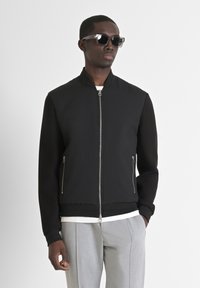 Giacca bomber nera con texture a coste sul davanti e maniche lisce, dotata di una cerniera frontale e tasche laterali. Indossata sopra una camicia bianca.