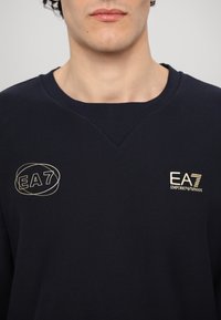 Jūras spēku džemperis, kas izgatavots no teksturēta auduma, ar V veida kakla izgriezumu un divām logo: viens zelta izšuvumā un otrs ovālā kontūrā.