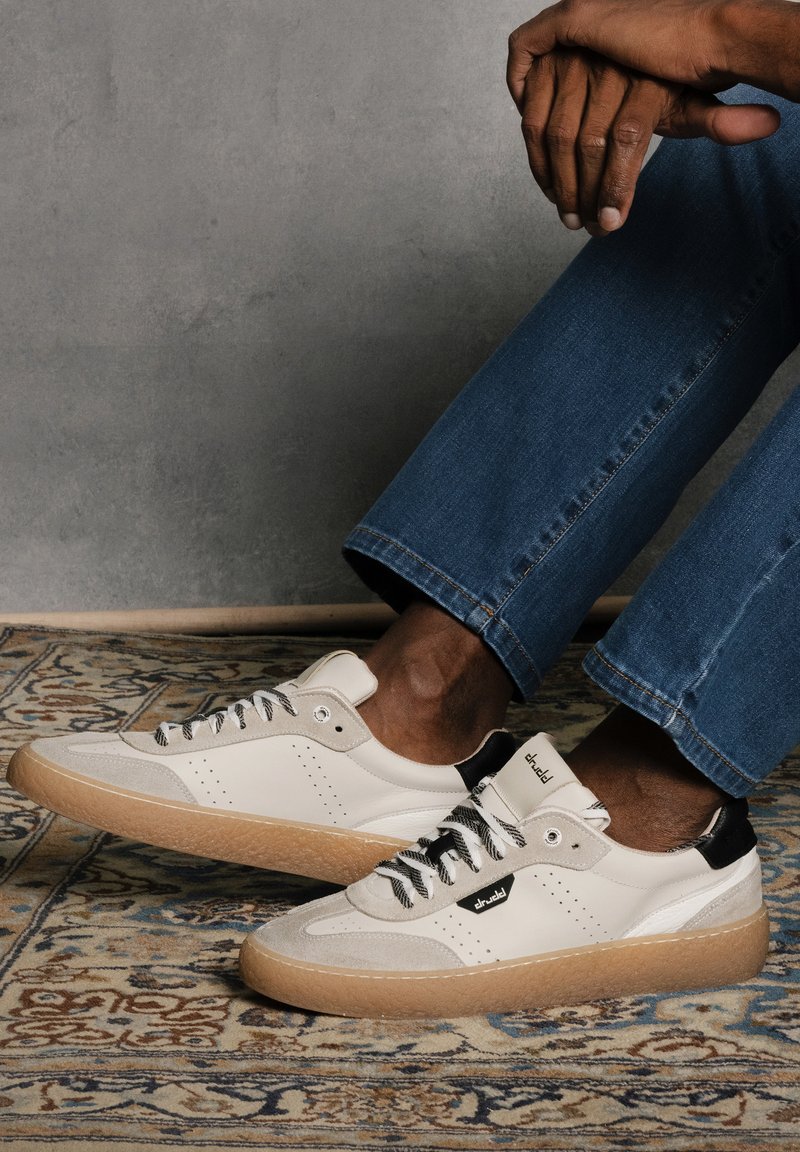 White sneakers with suede accents, black heel detail, and gray laces. Features perforations, a gum sole, and a textured upper design.