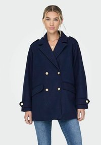 Manteau croisé bleu marine avec boutons dorés, grandes poches latérales et épaulettes ; fabriqué en tissu texturé.
