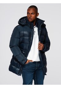 Hombre vestido con una chaqueta acolchada azul marino sobre una camisa polo blanca y jeans azules, con una mano en el bolsillo y mirando hacia un lado.