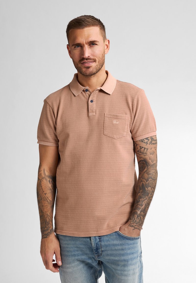 Polo triko v prachově růžové barvě, texturovaná látka, krátké rukávy, tříknoflíková légou, náprsní kapsa s logem, v kombinaci se světle modrými džíny.