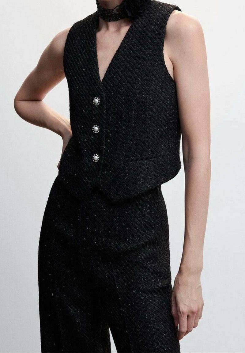 Veste noire sans manches texturée avec des boutons en perle, portée avec un pantalon taille haute assorti, sur une personne debout devant un fond uni.