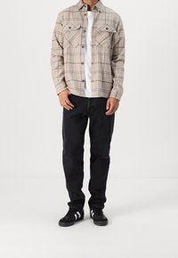 Beige flanellen overhemd met zwart geruit patroon, twee borstzakken, gedragen over een witte T-shirt, gecombineerd met zwarte jeans en zwarte sneakers.