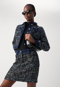 Ensemble de veste et jupe en tweed noir avec des paillettes bleues. Présente un design structuré, des boutons en métal et une coupe slim. Accessoires inclus : un sac à main noir.