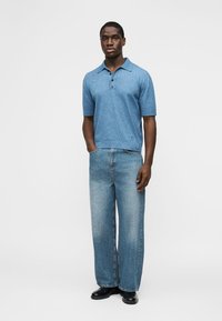 Homme debout, une main dans la poche, portant un polo bleu à manches courtes, un jean à jambes larges, et des chaussures noires sur un fond uni.