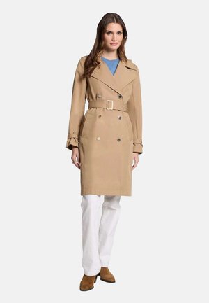 Trench-coat beige avec un devant à double boutonnage, taille ceinturée et grands revers. Présente des manches longues et des boutons, porté sur un pantalon blanc.