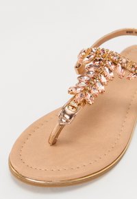 Sandale flip-flop bej cu un bandaj metalic auriu, împodobit cu strasuri roz și o cataramă decorativă. Talpă netedă, plată.