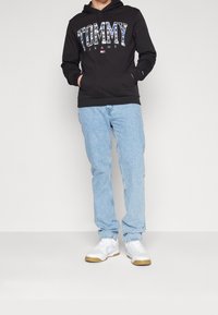Sudadera negra con logo blanco de "TOMMY JEANS", jeans de color azul claro y zapatillas blancas con suela de goma. La sudadera tiene un bolsillo frontal y cordón.