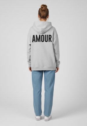 Felpa grigia con cappuccio e la scritta "AMOUR" stampata in nero sulla schiena. Abbinata a pantaloni leggeri a fantasia blu e sneakers bianche.