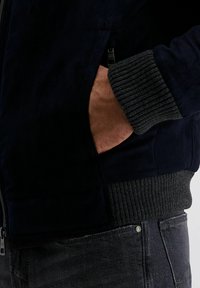 Marineblauwe suède jack met geribbelde donkergrauwe manchetten, zijzakken en een zichtbare rits. De stof heeft een gladde textuur.