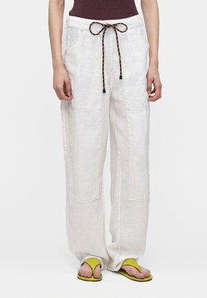 Pantalon blanc à jambes larges avec grandes poches avant et cordon marron, porté avec des sandales jaunes à anneau d’orteil et un haut sans manches foncé.
