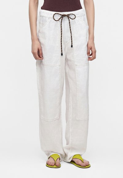 Pantalon blanc à jambes larges avec grandes poches avant et cordon marron, porté avec des sandales jaunes à anneau d’orteil et un haut sans manches foncé.