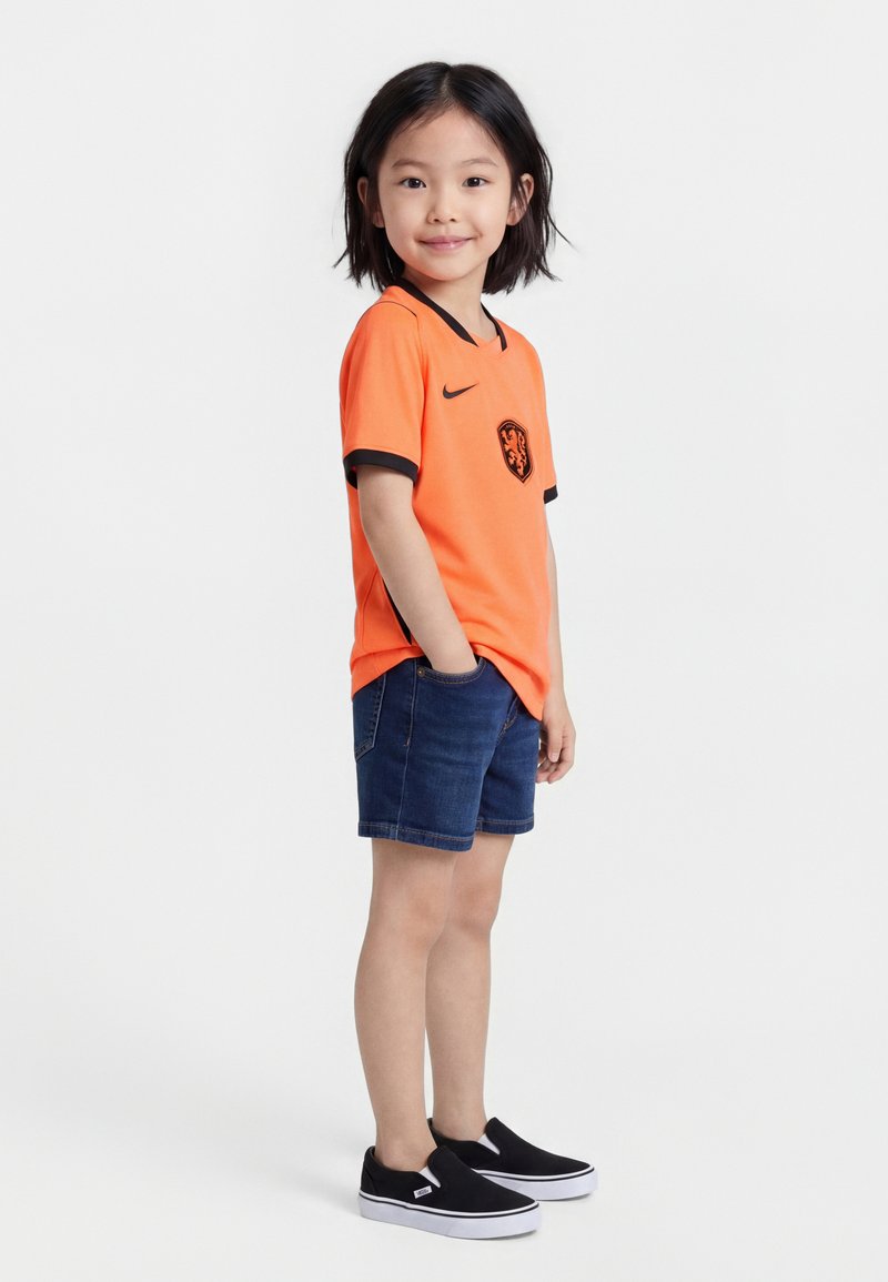 Νεαρό παιδί που στέκεται με τα χέρια στις τσέπες, φοράει πορτοκαλί αθλητική φανέλα, σκούρο τζιν σορτς και μαύρα παπούτσια χωρίς κορδόνια.