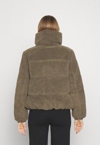 Mikina z fleecu v tlumené hnědé barvě s vysokým límcem, nadýchaným designem a elastickým pasem. Vyznačuje se texturovanou, měkkou látkou a prošitými detaily.