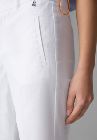Bílé kalhoty s hladkou strukturou, s jemnými bočními kapsami a stříbrným logem jako akcentem. U pasu je připevněna oranžová smyčka.