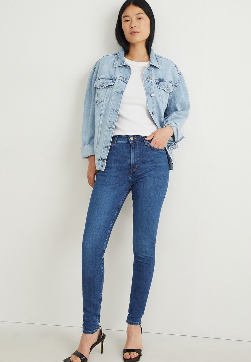 Eine Frau steht in einer blauen Jeansjacke, einem weißen T-Shirt, schmalen blauen Jeans und schwarzen offenen High Heels vor einem schlichten weißen Hintergrund.