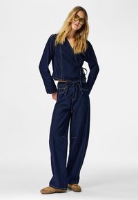 Tenue en denim composée d'une veste à enroulement avec manches longues et d'un pantalon à jambes larges. Les deux sont bleu foncé avec des coutures visibles et une ceinture nouée.