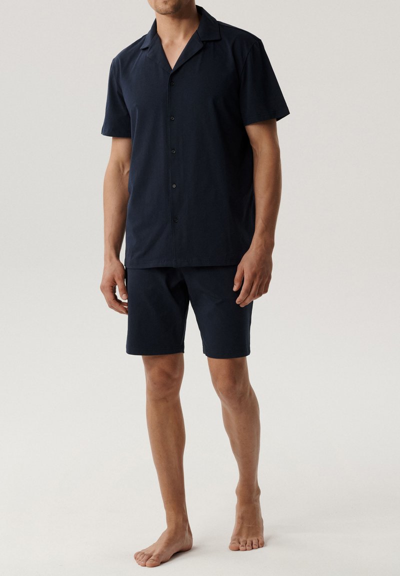 Granatowa, krótko rękawowa koszula zapinana na guziki z kołnierzykiem typu notch oraz pasujące szorty, wykonane z gładkiego materiału. Model boso stoi na neutralnym tle.
