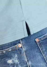 Lichtblauwe shirt met een zijsplit aan de onderkant, gecombineerd met versleten blauwe jeans met koperen accenten en een gestructureerde stof.