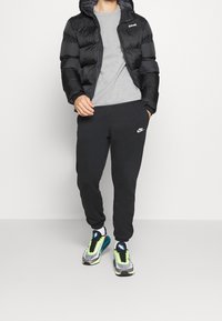 Giacca a vento nera con cappuccio, t-shirt grigia, pantaloni della tuta neri con logo Nike bianco e sneakers Nike multicolori con dettagli riflettenti.