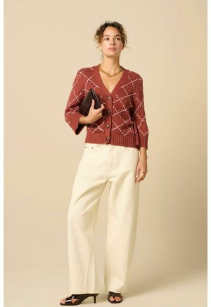 Frau trägt einen rostfarbenen gemusterten Cardigan, cremefarbene Hosen mit weitem Bein, schwarze Sandaletten mit Absatz, eine goldene Halskette und hält eine schwarze Clutch.