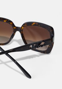 Ochelari de soare cu gradient maroniu, având o montură din scoică de țestoasă, cu un design pătrat masiv și un accent cu logo negru pe braț.
