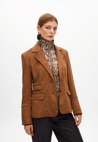 Hnědý semišový blejzr s přiléhavým střihem, dvěma předními kapsami a klopami, nošený přes vzorovaný top na zip.