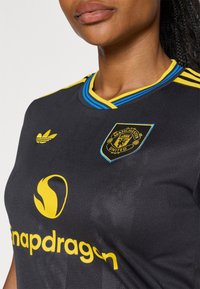 Melna sporta krekls ar dzelteniem un ziliem akcentiem. Iekļauts Adidas logo un Manchester United emblēma. Gluda tekstūra ar redzamām šuvju detaļām.
