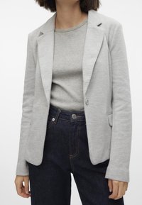 Světle šedý blazer vyrobený z měkké tkaniny s klopovými límci, jednořadým zapínáním na knoflík a dvěma předními kapsami, který je spárován se šedým žebrovaným topem a tmavými džíny.