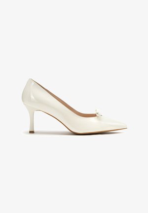 Zapato de tacón bajo de charol blanco y punta afilada con un pequeño lazo en el empeine. Textura suave y brillante con un diseño elegante.