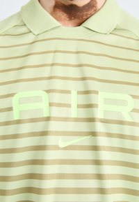Polo vert clair avec de fines rayures brunes. Présente un grand texte "AIR" en vert clair et un petit logo Nike dans la même couleur.