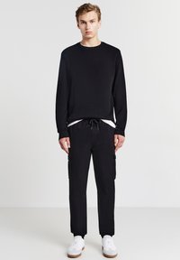 Junger Mann, der vor einem einfarbigen Hintergrund steht, trägt ein schwarzes Langarmshirt, schwarze Cargo-Hosen, weiße Sneakers und ein weißes Unterhemd.