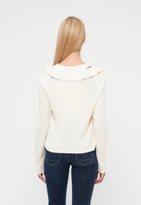 Πουλόβερ κρεμ χρώματος με γιακά με βολάν και μακριά μανίκια. Η πλάτη έχει λεπτομέρεια με κοψίματα, συνδυασμένο με σκουρόχρωμο μπλε τζιν.