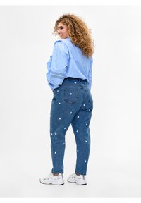 Gaiši zila blūze ar pūkainām piedurknēm un mežģīņu rotājumiem, saskaņota ar augsto jostas vietu džinsiem, uz kuriem ir balti sirdspati un brīvs piegriezums.