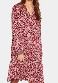 Punainen kukkamekko, jossa on V-mallinen kaula-aukko, pitkät hihat, kerätty vyötärö ja kerroksellinen helma. Kangas on koristeltu vaaleanpunaisilla ja valkoisilla terälehdillä.