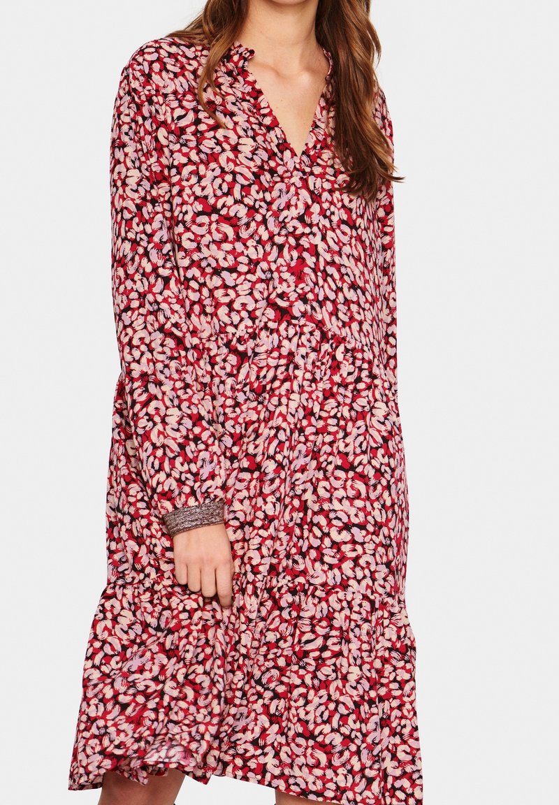 Punainen kukkamekko, jossa on V-mallinen kaula-aukko, pitkät hihat, kerätty vyötärö ja kerroksellinen helma. Kangas on koristeltu vaaleanpunaisilla ja valkoisilla terälehdillä.