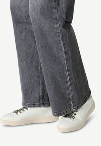 Sneakers bianchi con superficie texturizzata, suola beige e lacci decorativi, abbinati a jeans grigi scuri con taglio dritto e orlo leggermente sfilacciato.