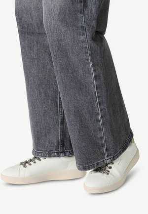 Sneakers bianchi con superficie texturizzata, suola beige e lacci decorativi, abbinati a jeans grigi scuri con taglio dritto e orlo leggermente sfilacciato.