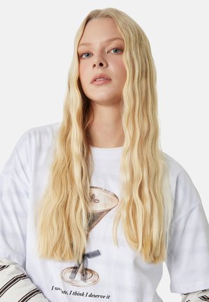 Joven mujer con cabello largo y rubio que lleva una camiseta gráfica blanca con una taza de café y mangas largas a rayas debajo.