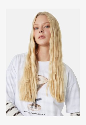 Junge Frau mit langen blonden Haaren trägt ein weißes T-Shirt mit einem Kaffee-Tassen-Motiv und darunter gestreifte lange Ärmel.