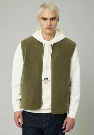 Gilet en polaire vert olive avec un col haut, porté sur un sweat à capuche blanc. Texture remarquable et design ouvert à l'avant, associé à un jean foncé.
