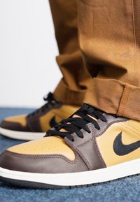 Brūni un dzelteni Nike sporta apavi ar melniem akcentiem, apaļām šņorēm un perforētu purngalu. Izstrādāti ar gludu un teksturētu materiālu kombināciju.