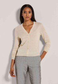 Frau mit langen dunklen Haaren, die einen beigefarbenen, geknöpften Cardigan und graue gestreifte Hosen trägt, steht vor einem schlichten, hellen Hintergrund.