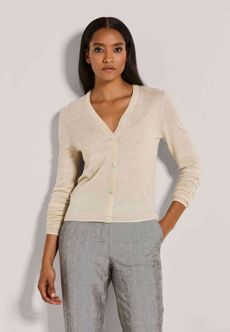 Frau mit langen dunklen Haaren, die einen beigefarbenen, geknöpften Cardigan und graue gestreifte Hosen trägt, steht vor einem schlichten, hellen Hintergrund.