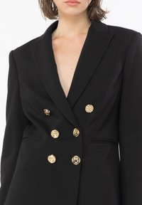 Blazer nero con scollo a V profondo, design doppiopetto e sei bottoni dorati; spalle strutturate e tessuto morbido.