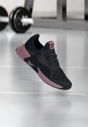 Czarno-różowy but sportowy Reebok unoszący się w pomieszczeniu, w tle rozmyte sprzęty siłowni na błyszczącej podłodze.