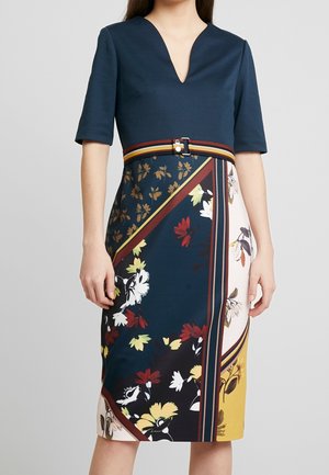 Kobieta ubrana w sukienkę do kolan z granatową, jednolita górą, dekoltem w serek, półdługimi rękawami, paskiem i wielokolorową spódnicą w kwiaty.